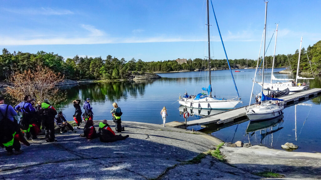 Grupp av människor instrueras innan segling. På klippa vid vatten med brygga och båtar